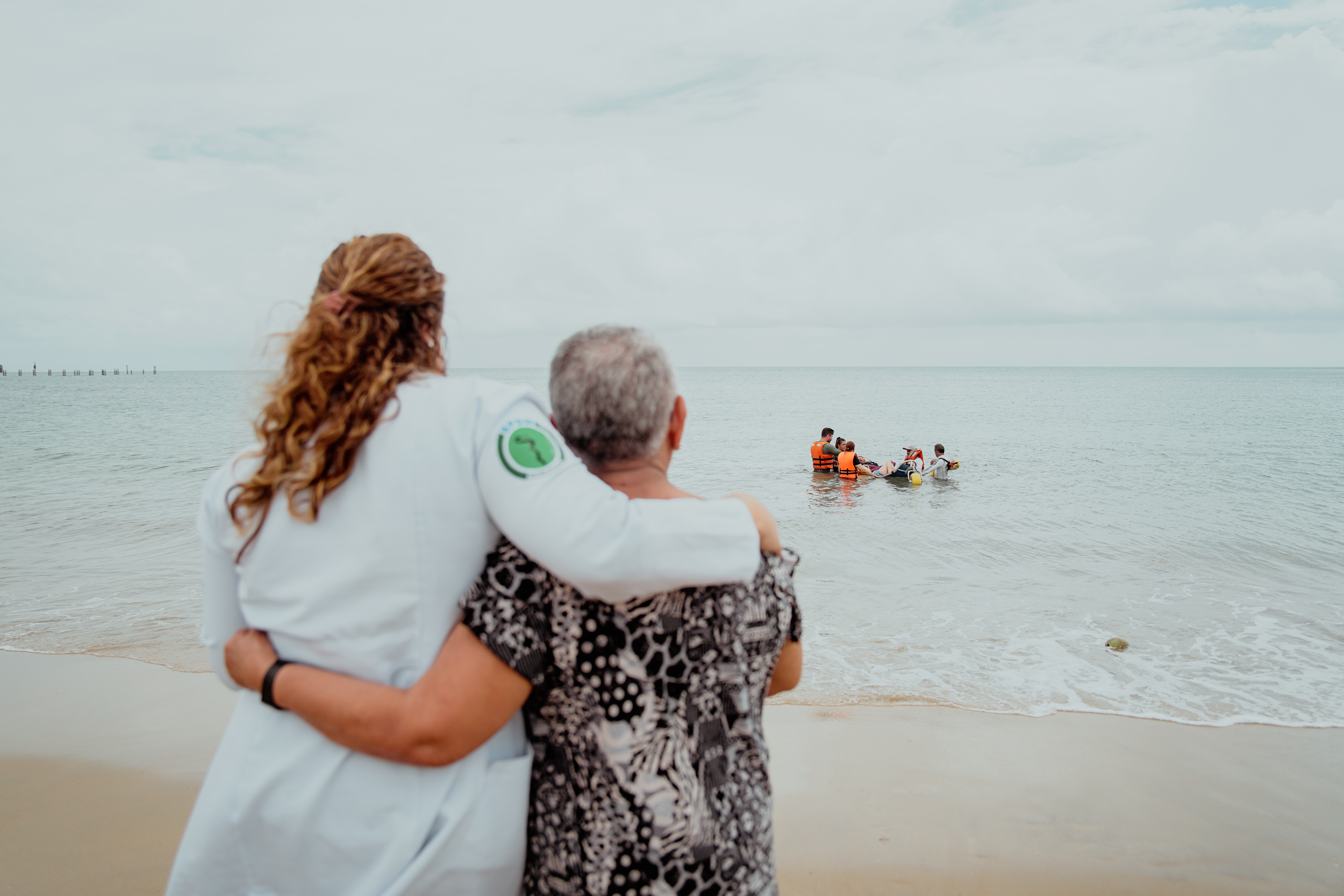 uma profissional de saúde e uma senhora observam da praia enquanto um jovem com paralisia cerebral toma banho de mar em veículos próprio e acompanhado por guarda-vidas